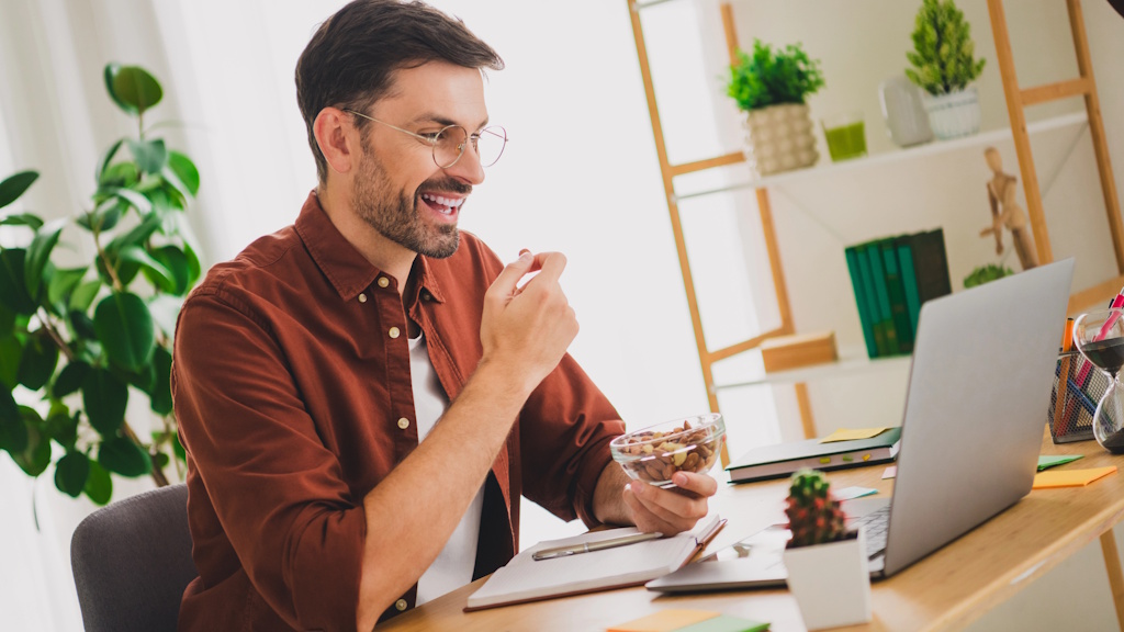 Selbstständiger Mann arbeitet am Laptop im Homeoffice und isst Nüsse als gesunden Snack für mehr Leistungsfähigkeit