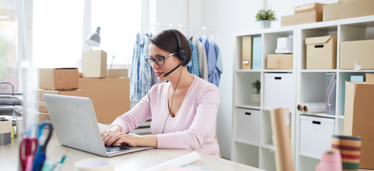 Frau mit Headset arbeitet am Laptop in einem hellen Büro mit Regalen, Kartons und Kleidung im Hintergrund