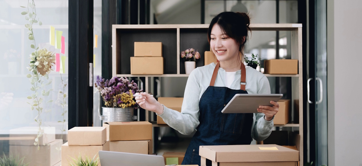 Frau mit Schürze organisiert Pakete und prüft Bestellungen per Tablet in einem kleinen Versand- oder Onlinehandelsbetrieb.