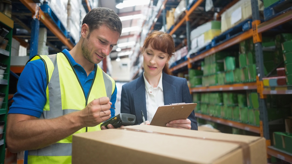 Lagerarbeiter scannt Paket im Hochregallager, während eine Mitarbeiterin die Daten auf einem Klemmbrett prüft
