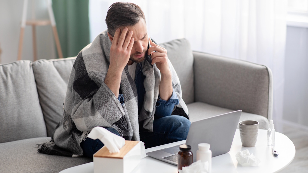 Die passende gesetzliche Krankenkasse für Selbstständige_TB Erkrankter Selbstständiger telefoniert zu Hause vor seinem Laptop – gesetzliche Krankenkasse für Selbstständige